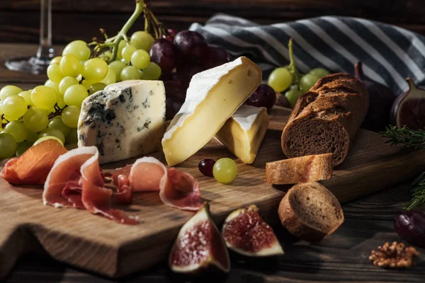 Diferentes tipos de queijos, presunto e uvas na tábua de corte — Fotografia de Stock