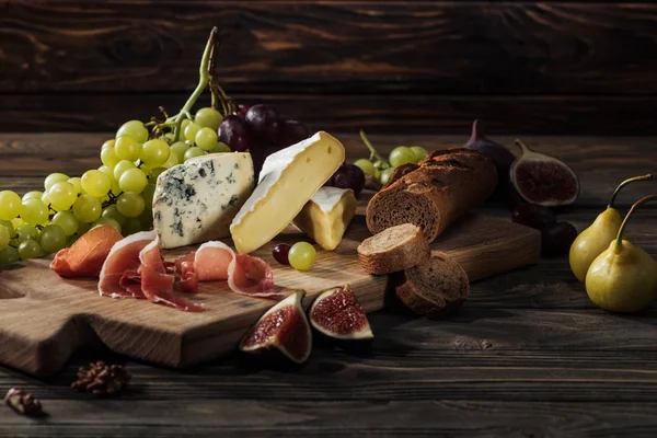 Different types of cheeses, prosciutto and baguette on cutting board — Stock Photo