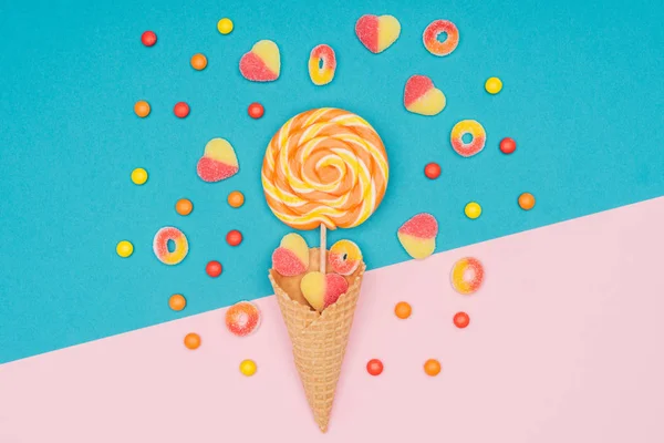 Vista superior de caramelos de gelatina, piruleta y cono de gofre en la superficie azul y rosa — Stock Photo