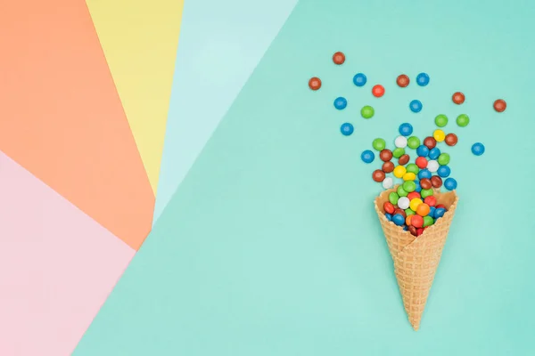 Вид сверху на разбросанные яркие драже конфеты и вафельный конус на цветной поверхности — стоковое фото