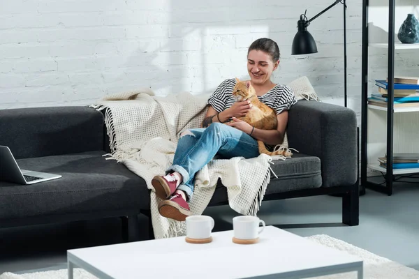 Bela menina sorridente sentado no sofá e palmando gato gengibre bonito na sala de estar — Fotografia de Stock