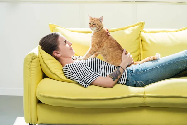 Menina bonita deitada no sofá amarelo e palmando bonito gato vermelho na sala de estar — Fotografia de Stock