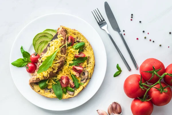 Flat lay with omelette with cherry tomatoes, avocado pieces and cutlery on white marble surface — Stock Photo