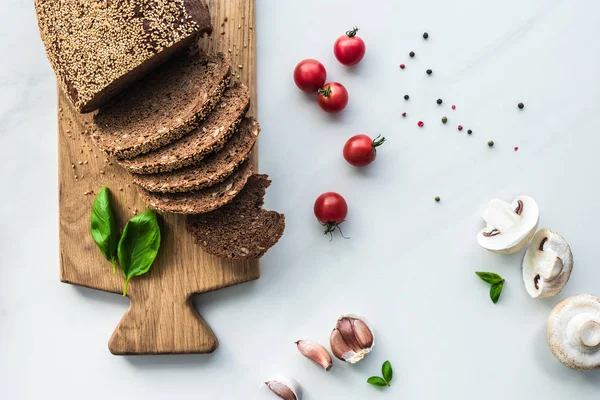 Tendido plano con pan cortado en tabla de cortar de madera, pimienta negra y verduras para cocinar el desayuno en la superficie de mármol blanco - foto de stock