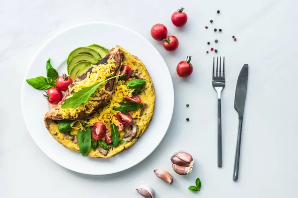 Top view of homemade omelette with cherry tomatoes, avocado pieces and cutlery on white marble surface — Stock Photo