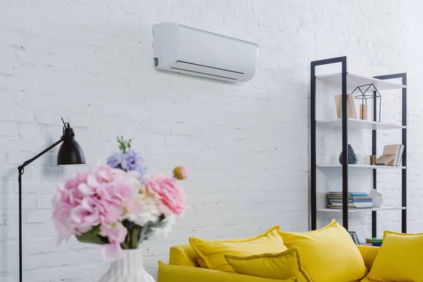 Интерьер современной гостиной с цветами в вазе, желтый диван и кондиционер висит на белой стене — стоковое фото