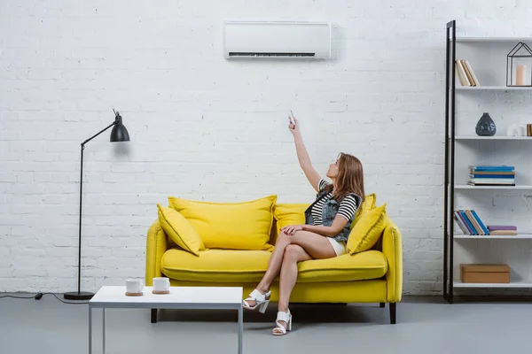 Attraktive junge Frau sitzt auf Sofa und zeigt auf Klimaanlage mit Fernbedienung — Stockfoto