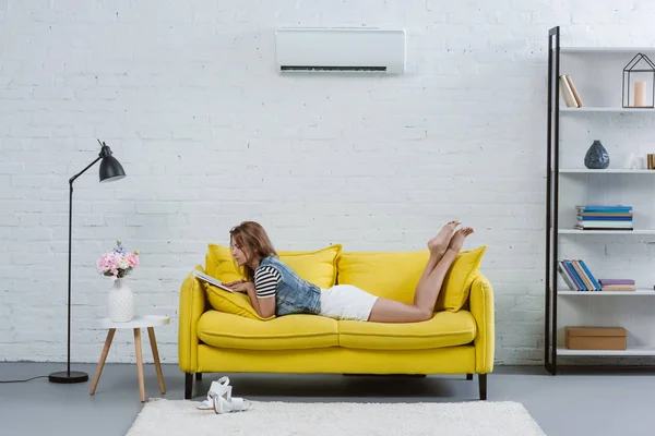Hermosa mujer joven lectura libro en sofá bajo el aire acondicionado colgando en la pared - foto de stock