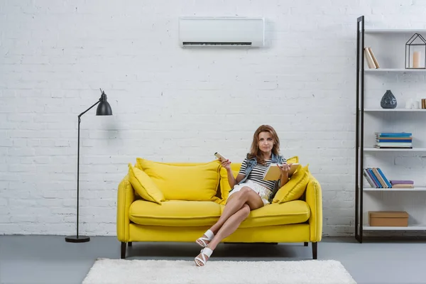 Focused young woman with book sitting on sofa and pointing at air conditioner with remote control — Stock Photo