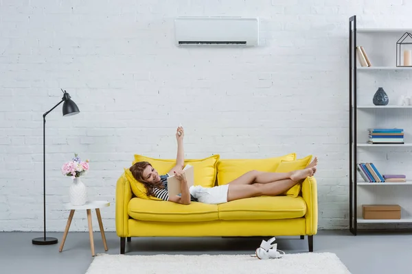 Mujer joven relajada leyendo libro en el sofá y apuntando al aire acondicionado con control remoto - foto de stock