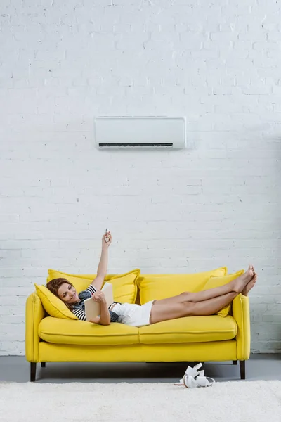 Glückliche junge Frau liest Buch auf Sofa und zeigt auf Klimaanlage mit Fernbedienung — Stockfoto