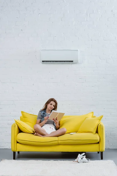 Красива молода жінка читає книгу на дивані під кондиціонером, що висить на стіні — стокове фото