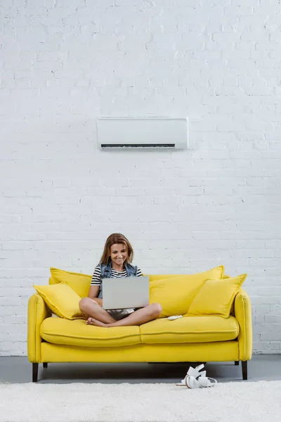 Приваблива молода жінка працює з ноутбуком, сидячи на дивані під кондиціонером, що звисає на стіні — стокове фото