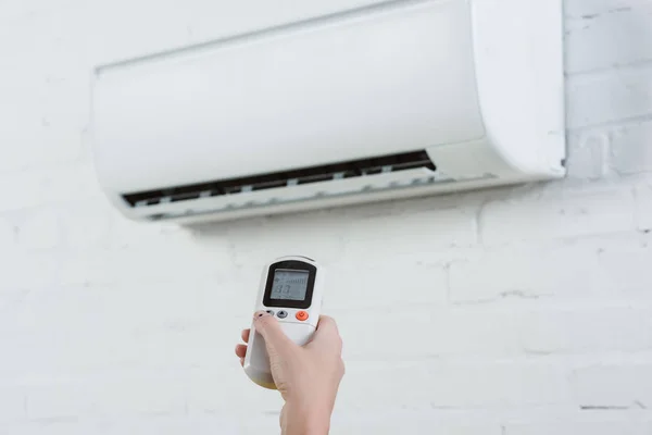 Cropped shot of woman pointing at air conditioner hanging on white brick wall with remote control — Stock Photo