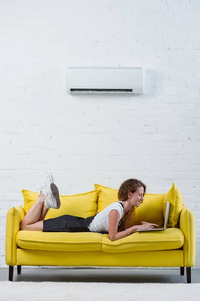 Счастливая молодая женщина работает с ноутбуком, лежа на диване под висящим на стене кондиционером — стоковое фото