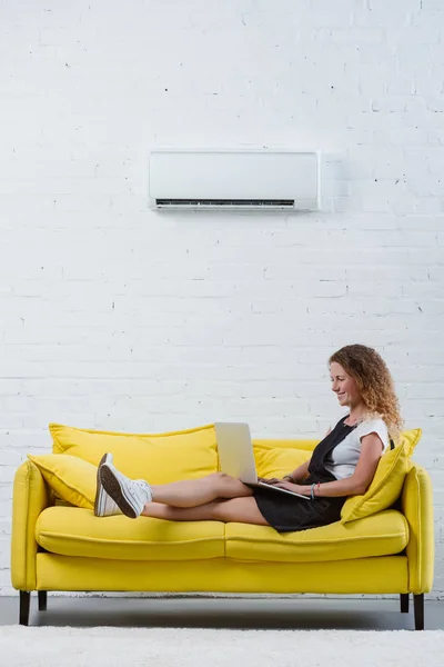 Щаслива молода жінка працює з ноутбуком, сидячи на дивані під кондиціонером, що звисає на стіні — стокове фото