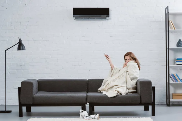 Mujer joven cubierta con manta en el sofá bajo el aire acondicionado colgando en la pared - foto de stock