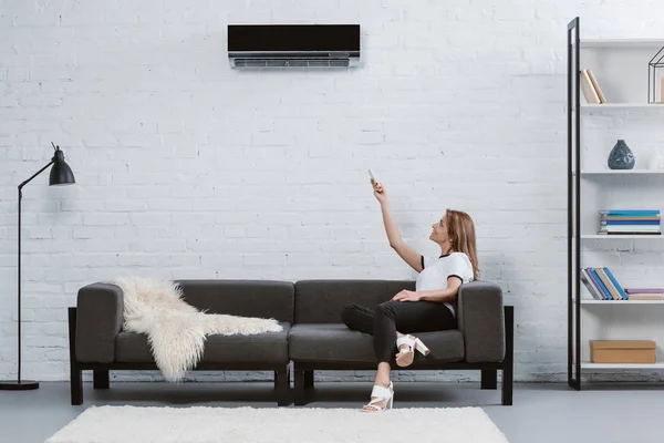 Schöne junge Frau sitzt auf Sofa und zeigt auf Klimaanlage, die mit Fernbedienung an der Wand hängt — Stockfoto