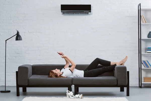 Feliz joven mujer usando teléfono inteligente en el sofá bajo el aire acondicionado colgando en la pared - foto de stock