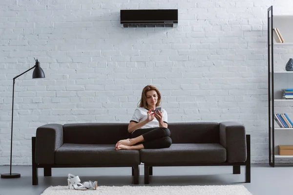 Mujer joven y relajada usando teléfono inteligente en el sofá bajo el aire acondicionado colgando en la pared - foto de stock