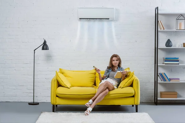 Beautiful young woman with book sitting on couch and pointing at air conditioner hanging on wall with remote control — Stock Photo