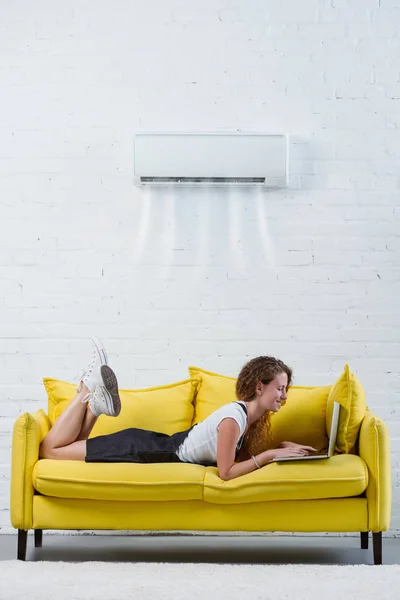 Atractiva mujer joven que trabaja con el ordenador portátil en el sofá bajo el aire acondicionado colgando en la pared - foto de stock