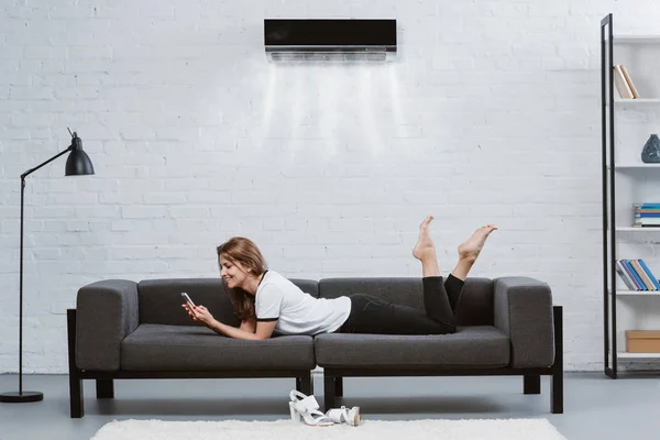 Щаслива молода жінка використовує смартфон на дивані під кондиціонером, що висить на стіні — стокове фото