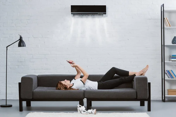 Mujer joven y relajada usando teléfono inteligente en el sofá bajo el aire acondicionado colgando en la pared - foto de stock