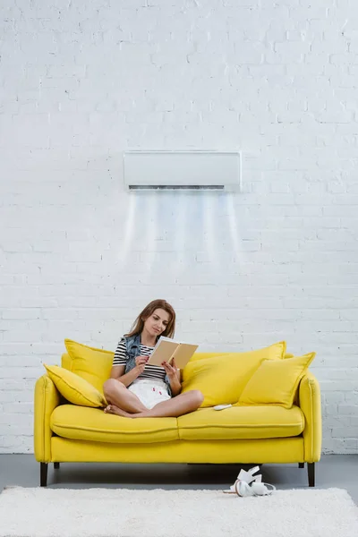 Centrado libro de lectura de la mujer joven en el sofá bajo el aire acondicionado colgando en la pared y soplando aire fresco - foto de stock