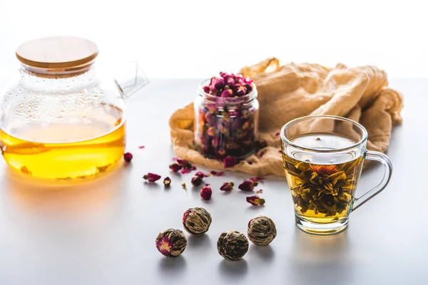 Capullos de rosas secas en frasco, taza de té floreciente chino con bolas de té en la mesa blanca - foto de stock