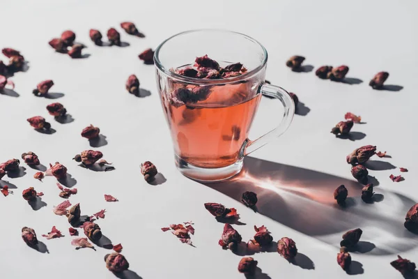 Chá de botões de rosa seco na xícara e rosas espalhadas na mesa branca — Fotografia de Stock