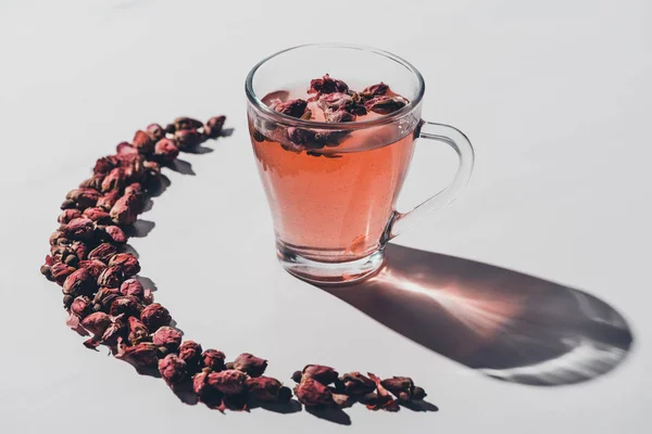 Сушеные бутоны роз чай в чашке и отражающая тень на белом столе — стоковое фото
