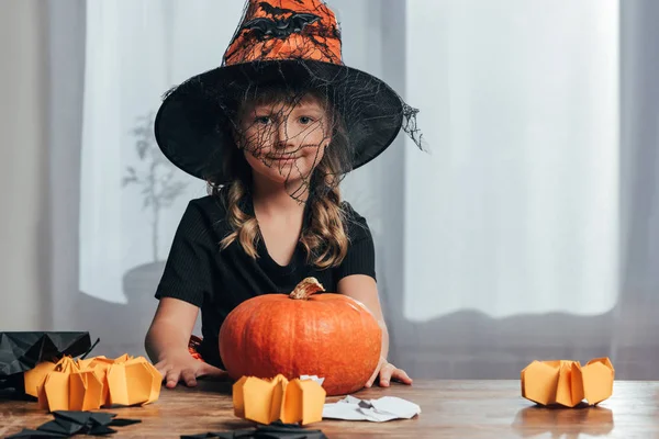 Retrato de adorable niño en traje de Halloween bruja sentado en la mesa con calabaza en casa - foto de stock