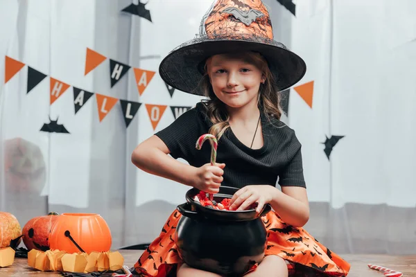 Portrait of smiling child in witch halloween costume with black pot full of candies at home — Stock Photo