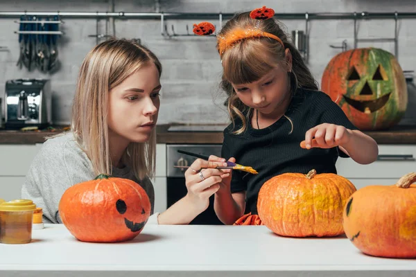 Mujer joven y hermana pequeña pintando calabazas para Halloween juntos en la cocina en casa - foto de stock