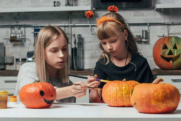 Mujer joven y hermana pequeña pintando calabazas para Halloween juntos en la cocina en casa - foto de stock