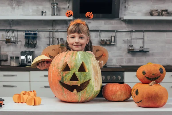 Retrato de niño sonriente en traje de halloween de pie en la calabaza tallada grande en la mesa en la cocina en casa - foto de stock