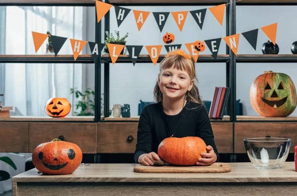 Portrait of smiling kid at tabletop with pumpkins and hanging happy halloween flags behind at home — Stock Photo