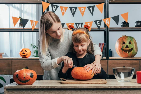 Joven mujer ayudando a hermana pequeña en halloween traje tallado calabazas en casa - foto de stock