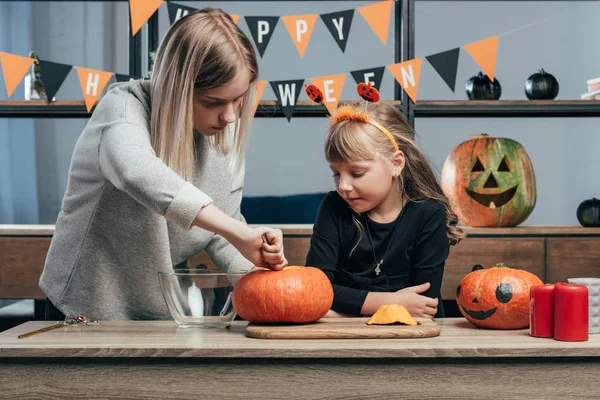 Joven mujer ayudando a hermana pequeña en halloween traje tallado calabazas en casa - foto de stock