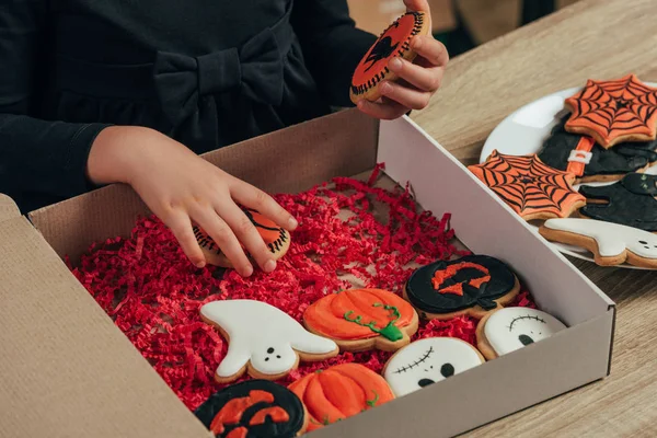 Teilansicht eines Kindes, das zu Hause an einer hölzernen Tischplatte thematische Halloween-Plätzchen arrangiert — Stockfoto