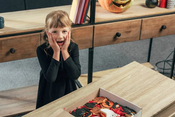 High angle view of shocked kid looking at camera at tabletop with halloween cookies in box at home — Stock Photo