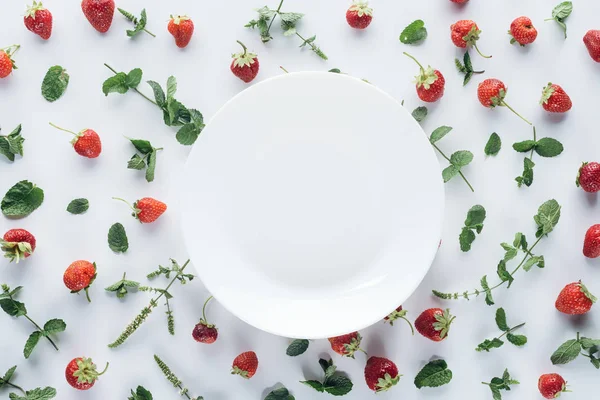 Вид сверху на пустую тарелку в окружении спелых клубничных и мятных листьев на белой столешнице — стоковое фото