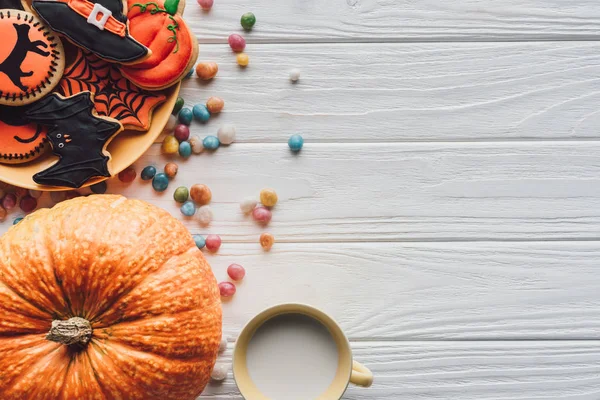 Vista elevada de calabaza, plato con galletas de halloween, caramelos y taza con leche sobre fondo de madera - foto de stock