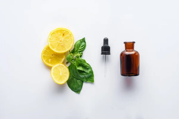 Vista superior da garrafa de óleo essencial de ervas naturais, conta-gotas, pedaços de limão e folhas verdes isoladas em branco — Fotografia de Stock