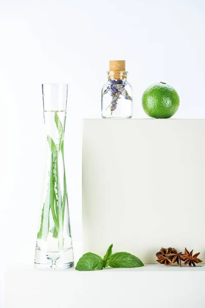 Botella transparente y jarrón de aceites esenciales de hierbas naturales, cal y clavel en la superficie blanca - foto de stock