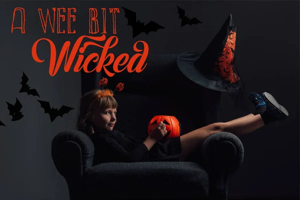 Adorable kid in halloween costume resting in armchair in dark room with 