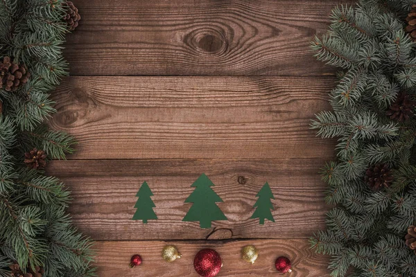 Draufsicht auf grüne Papiertannen mit glänzenden Kugeln und Nadelzweigen mit Tannenzapfen auf Holzoberfläche, weihnachtlicher Hintergrund — Stockfoto