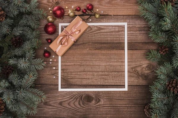 Vue de dessus du cadre blanc, du cadeau de Noël et des boules brillantes sur la surface en bois avec des rameaux de sapin et des cônes de pin — Photo de stock