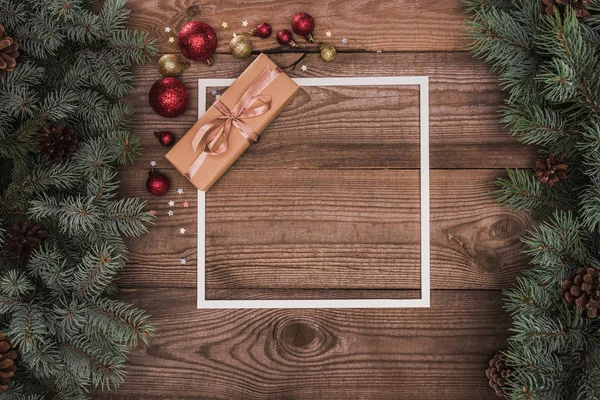 Top view of white frame, christmas present and shiny baubles on wooden surface with fir twigs and pine cones — Stock Photo